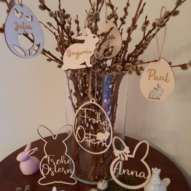 Verzierte Holzanhänger mit Namen und Ostermotiven an einer Vase mit Zweigen.